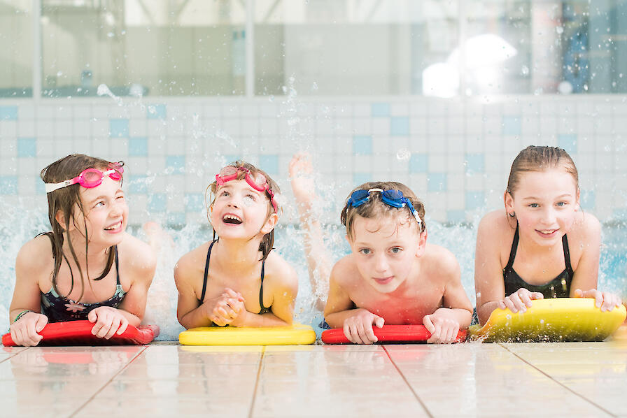 Vier Kinder liegen am Schwimmbeckenrand auf Schwimmbrettern und haben Spaß.