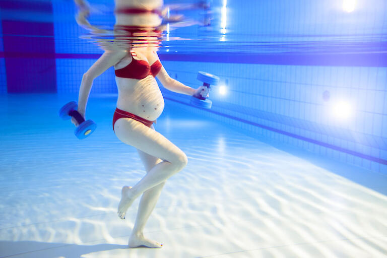 Unterwasserfoto von einer schwangeren Frau im roten Bikini mit zwei AquaFitness-Hanteln in der Hand.