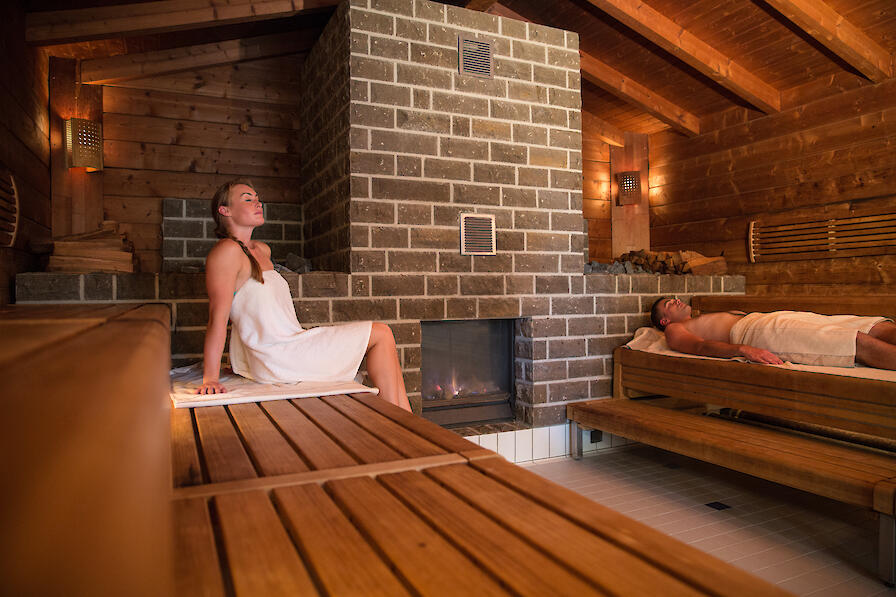 Kaminsauna Takka mit einem großen Kamin sowie einem Mann und einer Frau, die sich in der Sauna entspannen.