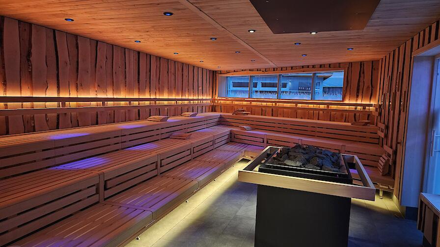 Beleuchtete Aufguss-Sauna „Löyly“ mit einem Saunaofen in der Mitte und Fenstern in den Saunagarten.