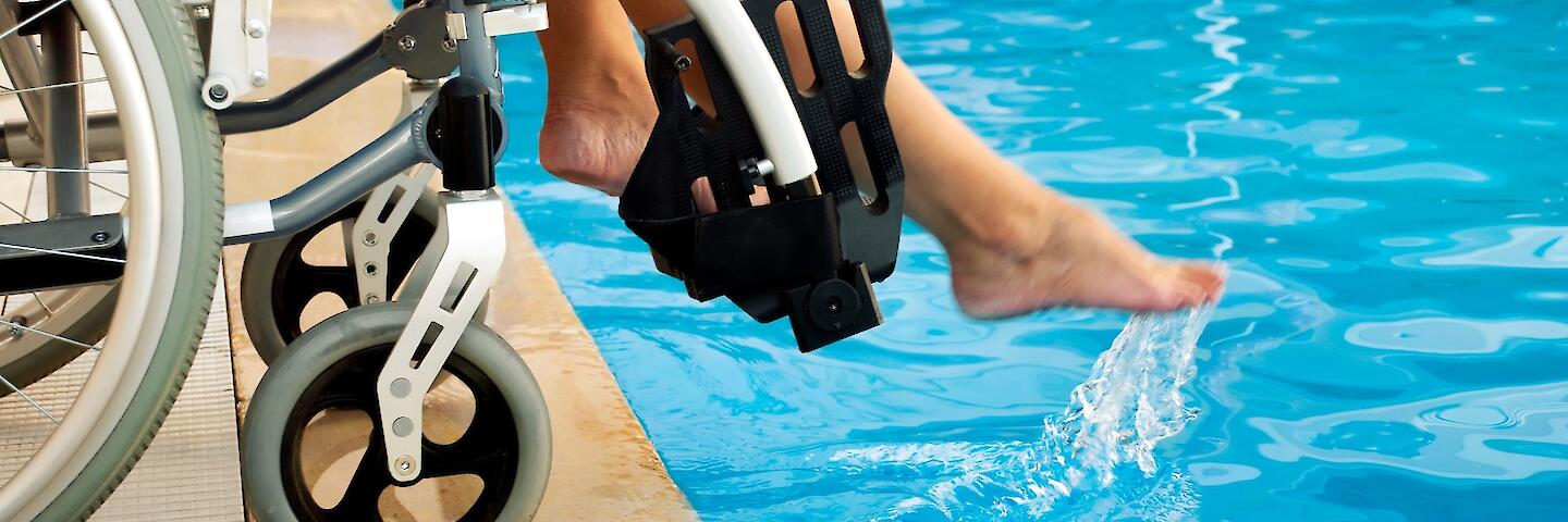 Ein Rollstuhl steht an dem Rand eines Schwimmbeckens und die Person im Rollstuhl hält ein Fuß ins Wasser.