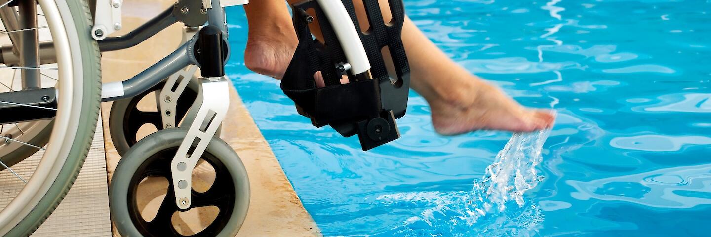 Ein Rollstuhl steht an dem Rand eines Schwimmbeckens und die Person im Rollstuhl hält ein Fuß ins Wasser.