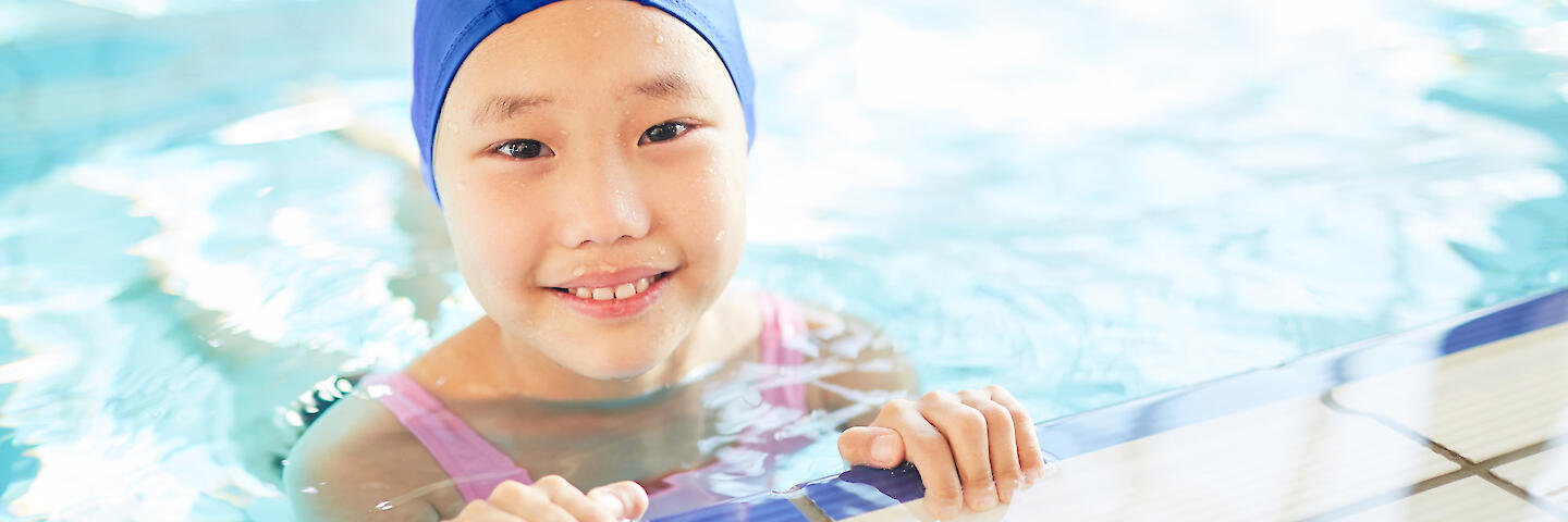 Glückliches Mädchen mit blauer Badekappe ist im Schwimmbecken und hält sich am Beckenrand fest.