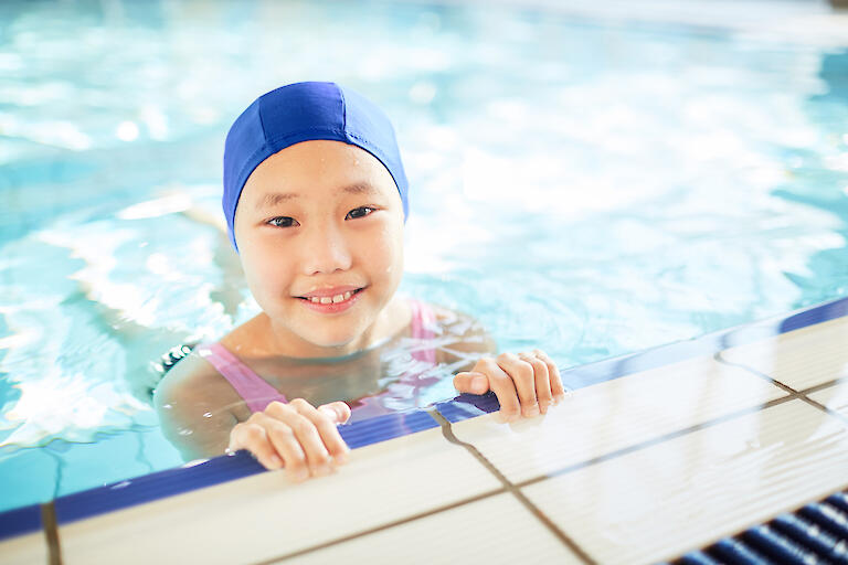 Glückliches Mädchen mit blauer Badekappe ist im Schwimmbecken und hält sich am Beckenrand fest.