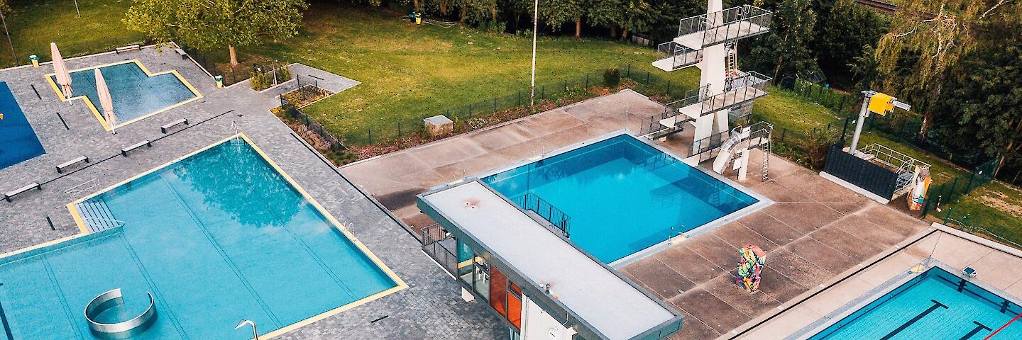 Luftansicht des Freibades mit dem 50-Meter-Becken, dem Sprungturm, dem Nichtschwimmerbecken mit verschiedenen Attraktionen, dem Kinderplanschbecken sowie dem Wasserspielplatz.