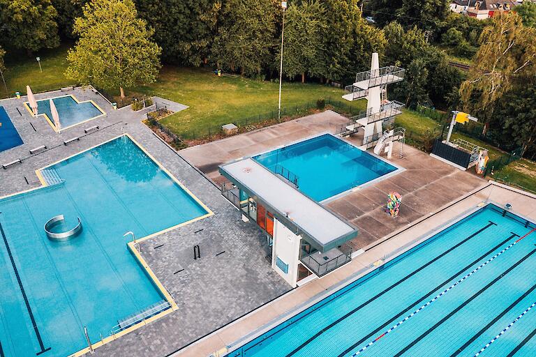 Luftansicht des Freibades mit dem 50-Meter-Becken, dem Sprungturm, dem Nichtschwimmerbecken mit verschiedenen Attraktionen, dem Kinderplanschbecken sowie dem Wasserspielplatz.