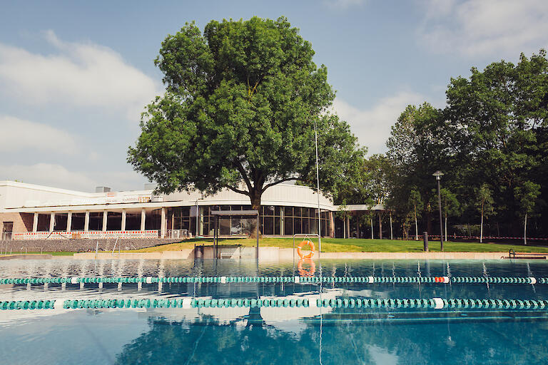 Der Bereich des Mehrzweckbeckens, in dem Leinen gespannt sind um das Schwimmen auf 50m-Bahnen zu ermöglichen. Im Hintergrund das Gebäude des Hallenbades.